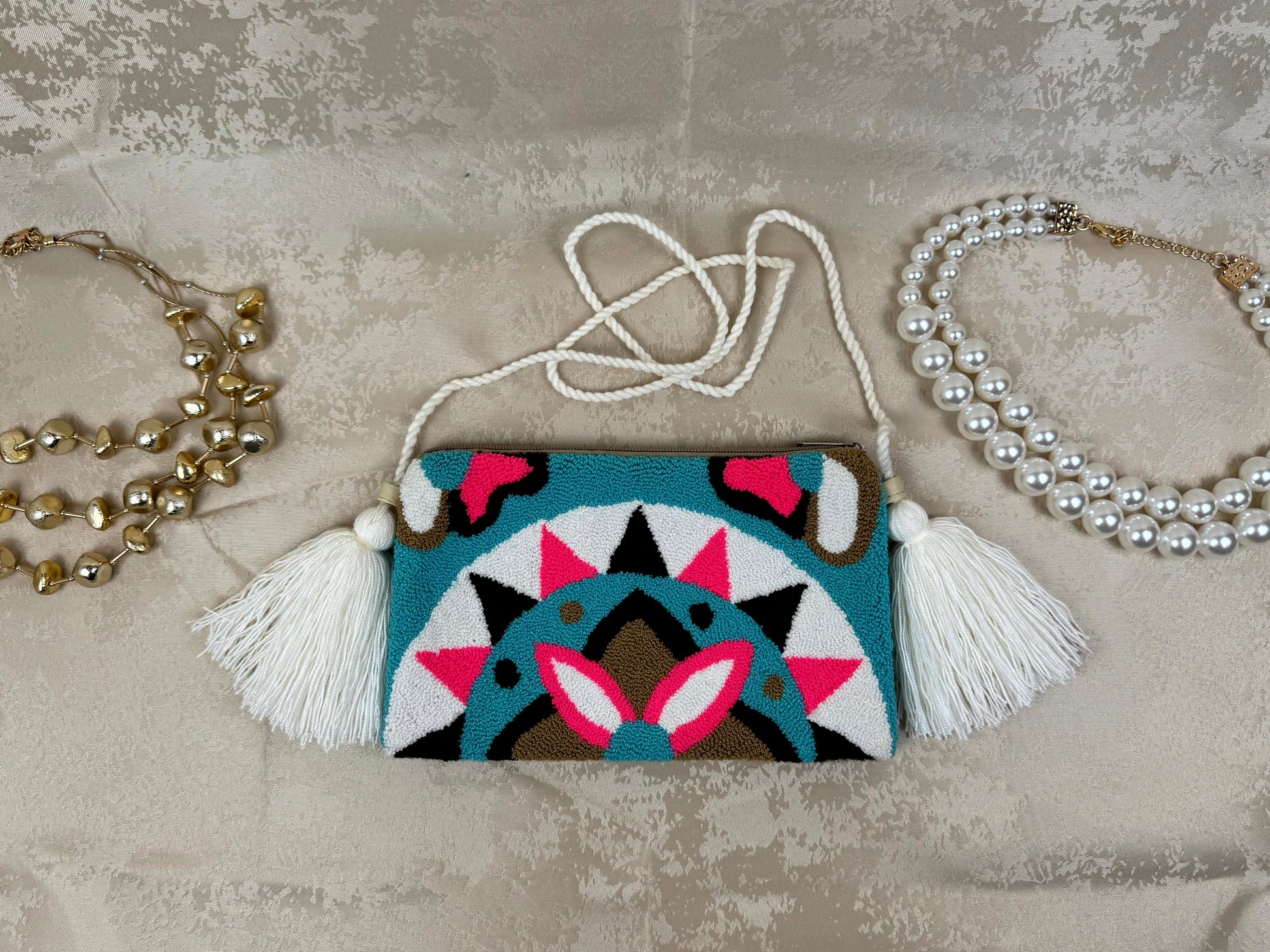 Small handbag with geometric design and tassels on a textured surface with two necklaces.